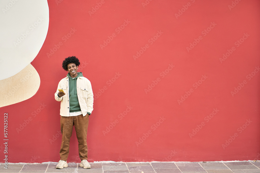 Happy smiling cool generation z young African American ethnic stylish hipster guy model standing at red city urban wall outdoors using cell phone mobile device, looking at camera, holding cellphone. - obrazy, fototapety, plakaty 
