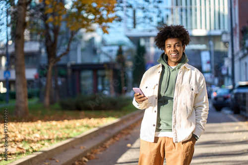 Smiling young African American guy user holding mobile phone standing with smartphone at sunny city street. Happy cool stylish hipster gen z teen using cell tech device outdoors, looking at camera.