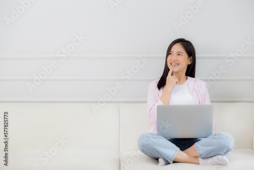Young asian woman working on laptop computer sitting on sofa in the living room at home, female is freelance sitting on couch using notebook and doubt about decision, one person, lifestyle concept.
