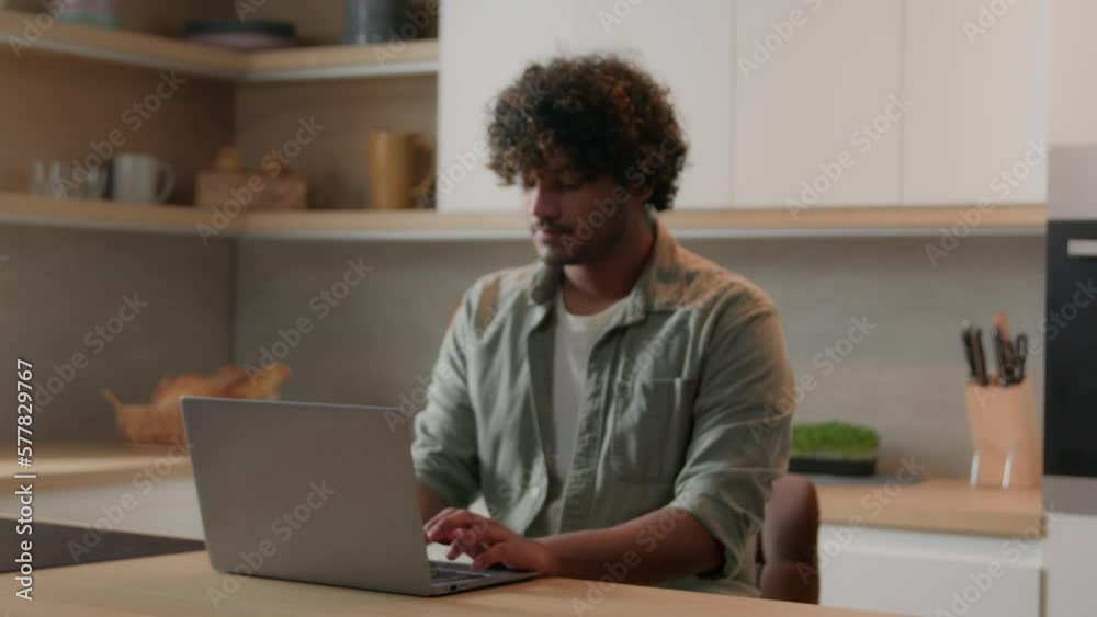 Moving shot focus from blurred to focused Indian business man working with laptop at kitchen smiling to camera freelancer Arabian computer distance work at table. Guy student studying online at home Moving shot focus from blurred to focused Indian business man working with laptop at kitchen smiling to camera freelancer Arabian computer distance work at table. Guy student studying online at home
