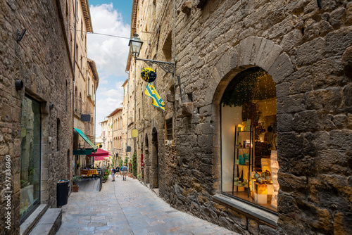 Tourists walk down a narrow cobblestone alley past shops and cafes in the historic medieval old town of the walled Tuscan city of Volterra, Italy. photo