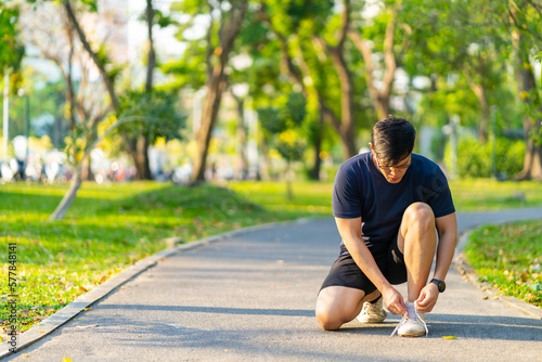 Asian man in sportswear tying running shoelaces during jogging exercise at public park in summer morning. Healthy guy athlete enjoy outdoor lifestyle sport training fitness workout running in the city