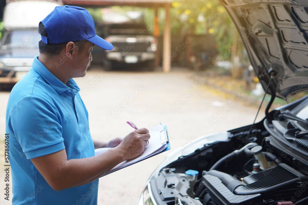 Asian man mechanic wears blue cap and blue shirt, holds paper, checking and analyzing car engine under the hood. Concept, Outdoor car inspection service. Claim for accident insurance. 