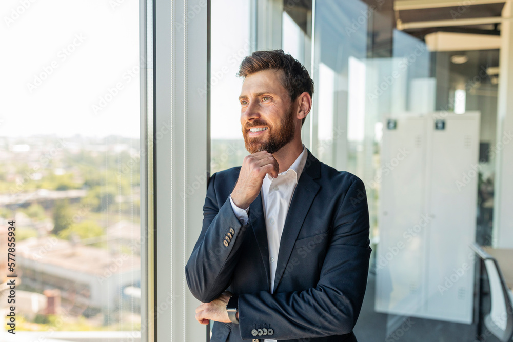 Happy confident businessman in suit standing near window in modern coworking office, waiting for meeting, free space Happy confident businessman in suit standing near window in modern coworking office, waiting for meeting, free space