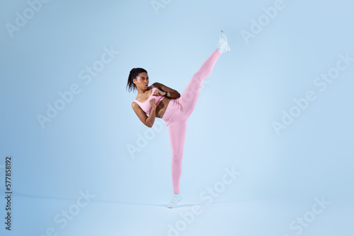 Determined black woman in sportswear doing high kick, tae-bo martial arts exercises on blue background, free space © Prostock-studio