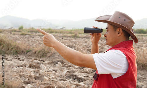 Asian man explorer wears hat, red vest shirt, holds binocular to explore boundary , points to something. Concept, land property exploration. Long vision distance, zoom for clearing scene. Observation