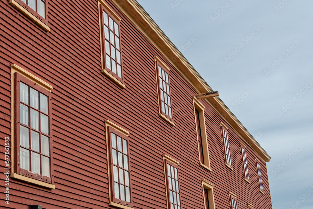 The exterior of a rusty red colored vintage warehouse with a wooden clapboard horizontal siding covered wall. There are two rows of windows with yellow trim, multi-glass panes, and a flat roof. 