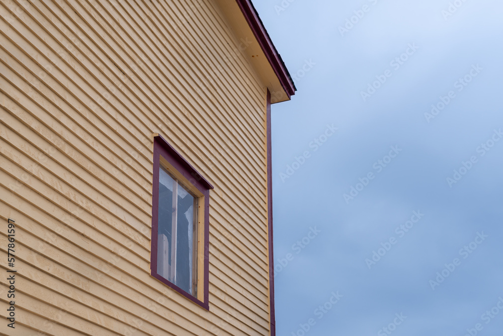 The exterior corner of an old wooden building. The wood clapboard walls are yellow in color with brown trim. There's a glass casement window with brown trim. A blue cloudy sky is in the background.