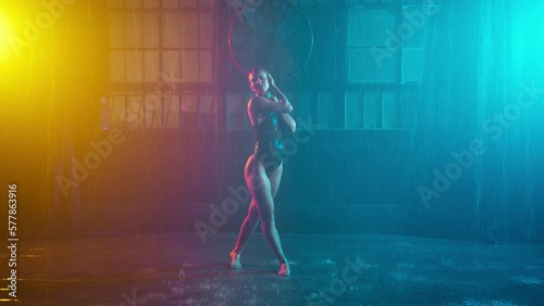 Hot dancer sitting on wet floor under rain garage background, moving sexually making erotic show teal orange spotlights. Seductive woman showing sexy body performing striptease cyberpunk urban stage