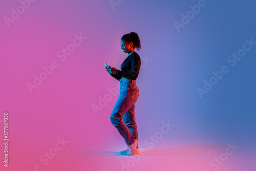 Fotografia Happy african american woman texting on cellphone, standing in colorful neon lig