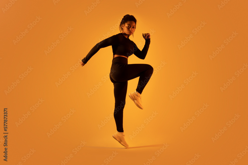 Sporty black woman jumping and doing stretching exercise over orange neon light background, full length