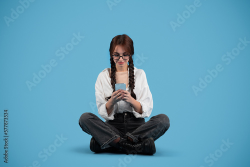 Smiling european teenager girl student with pigtails in glasses sits on floor, typing on phone isolated on blue studio background. Device for study, knowledge and education, communication remotely