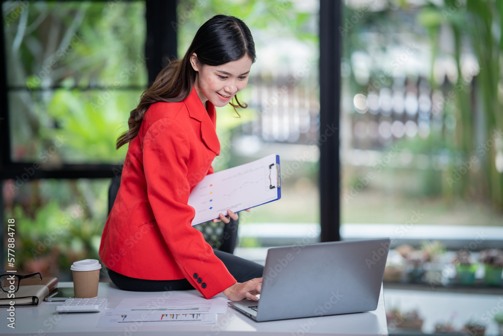 Attractive young smiling Asian woman in smart casual wear and holding smart phone looking at camera while leaning on desk in creative office Confident business expert Attractive young smiling Asian woman in smart casual wear and holding smart phone looking at camera while leaning on desk in creative office Confident business expert
