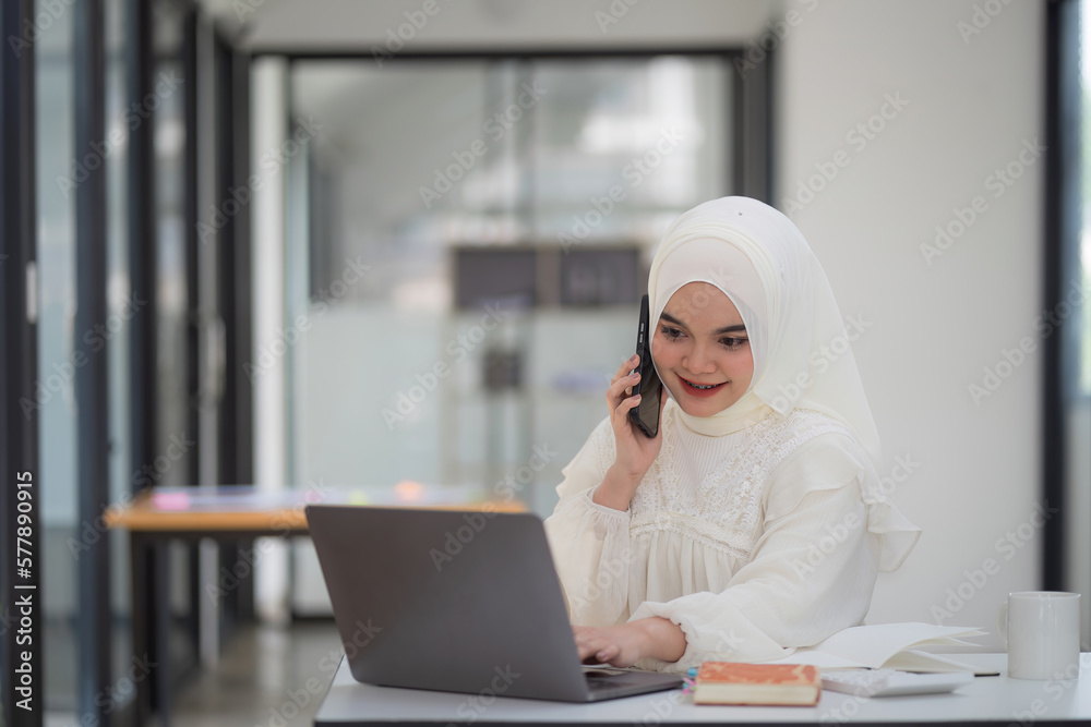 A happy and beautiful Muslim businesswoman in a white hijab is using her laptop at a cafe while engaging in a business discussion on her mobile phone.