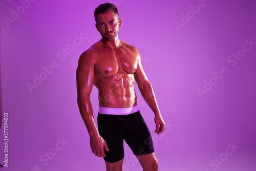 Man athletic body bodybuilder in briefs with nude torso abs full-length in the background fitness classes, purple colored light. Advertising, sports, active lifestyle, competition, challenge concept. 