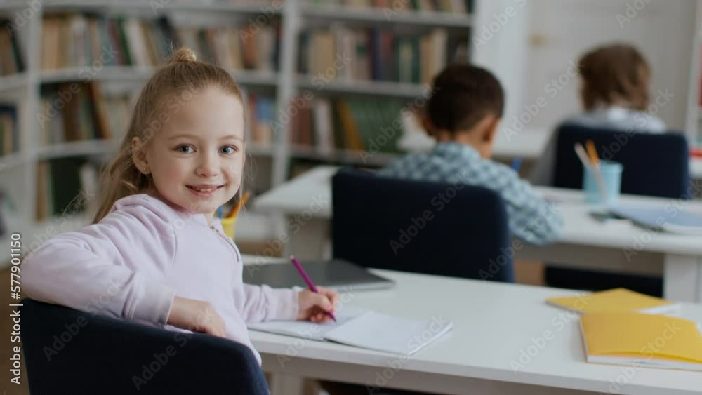Back view shot of group of little kids studying at classroom, cute schoolgirl turning to camera and smiling, empty space Back view shot of group of little kids studying at classroom, cute schoolgirl turning to camera and smiling, empty space