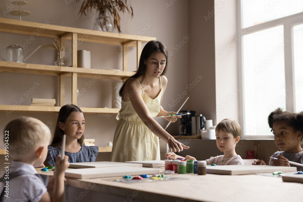 Pretty young artist woman teaching diverse group of little kids in artistic school studio. Children drawing pictures on class in acrylic, oil paints with assistance of female teacher Pretty young artist woman teaching diverse group of little kids in artistic school studio. Children drawing pictures on class in acrylic, oil paints with assistance of female teacher