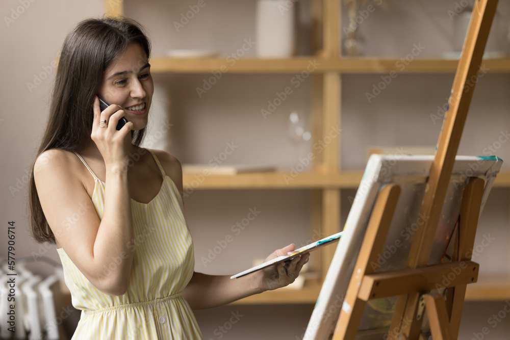 Happy positive young artist woman speaking on cell phone at easel, enjoying phone conversation, communication in art school studio, consulting teacher about drawing artwork