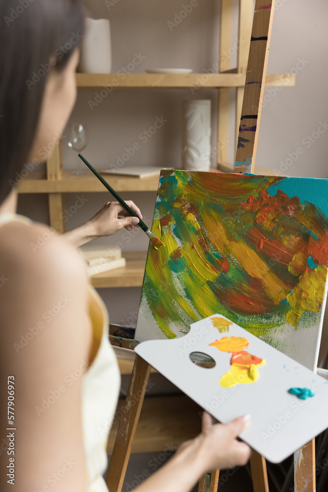 Young black haired artist woman practicing art therapy, drawing abstract colorful picture, painting in acryl on canvas on artistic class, holding palette, using brush, easel. Vertical shot