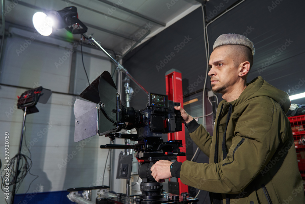 Serious caucasian videographer adjusts the composition on a professional movie camera on the set. a monoblock lighting fixture shines Serious caucasian videographer adjusts the composition on a professional movie camera on the set. a monoblock lighting fixture shines