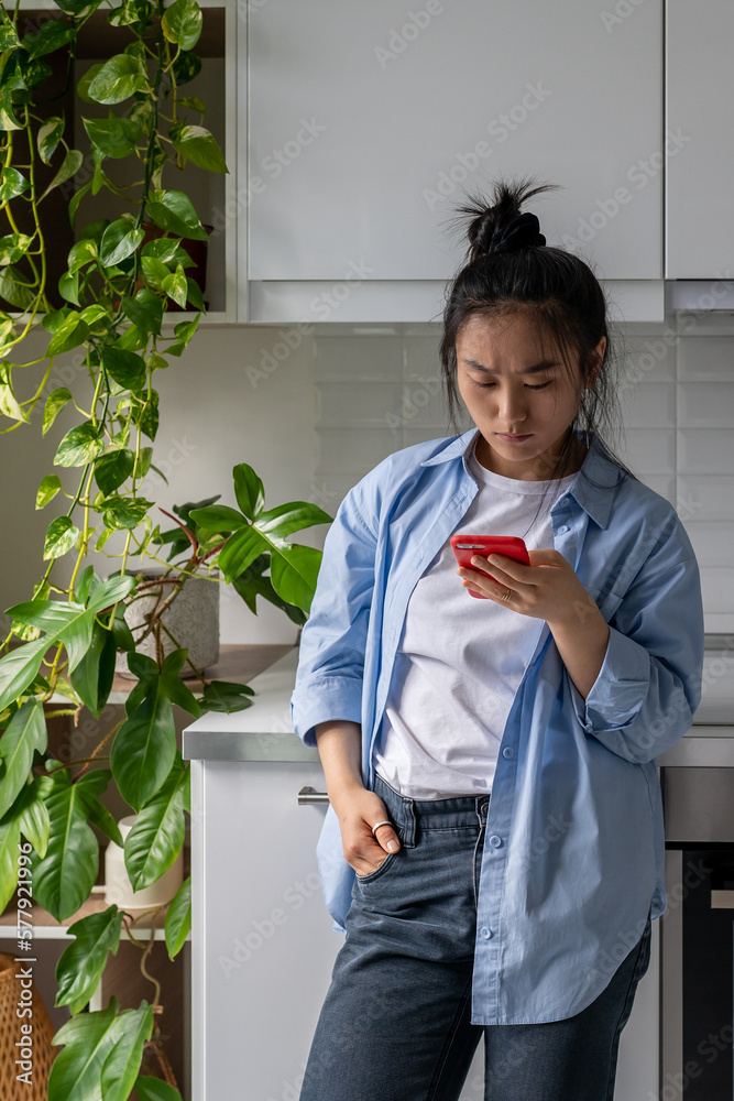 Serious Asian woman standing in kitchen at home holding mobile phone, reading bad news, receiving breakup message. Millennial girl looking at smartphone screen spying on boyfriend in social media Serious Asian woman standing in kitchen at home holding mobile phone, reading bad news, receiving breakup message. Millennial girl looking at smartphone screen spying on boyfriend in social media