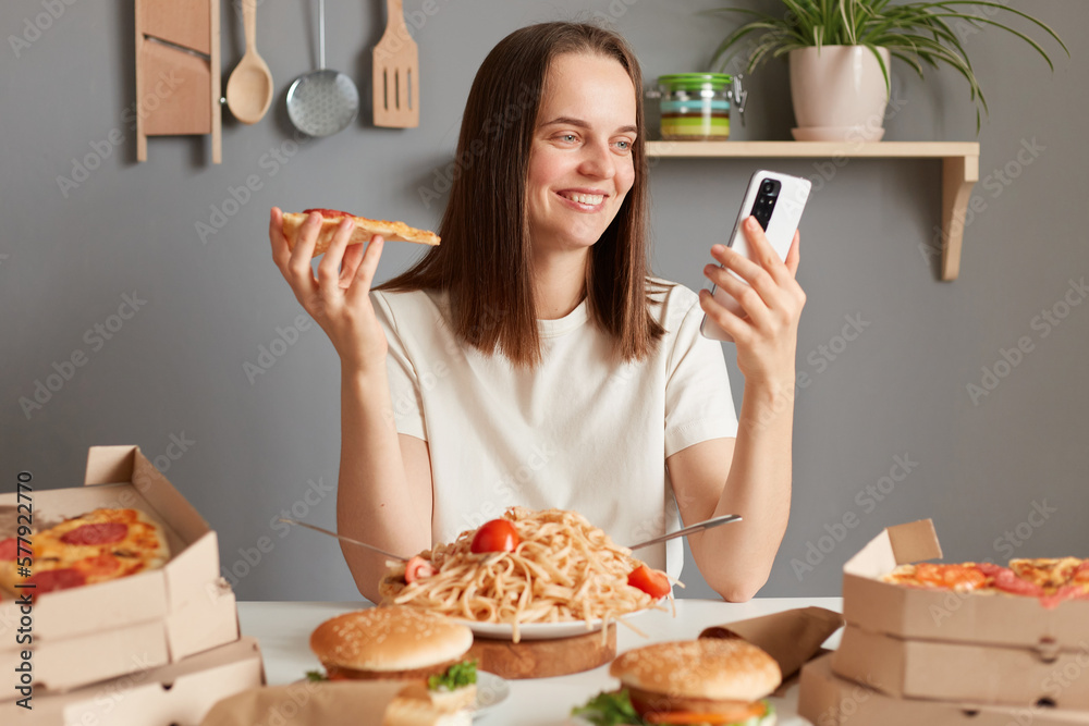 Photo of cheerful Caucasian young adult woman with brown hair wearing white t shirt sitting in kitchen at table and looking at smart phone display, having delicious snack, eating pizza.
