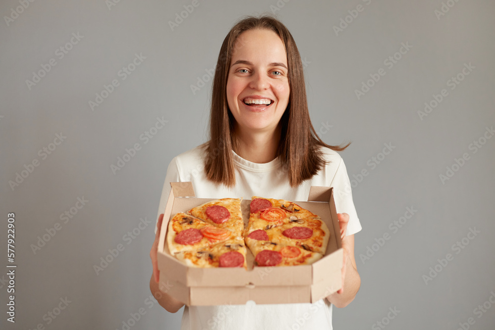 Portrait of attractive smiling positive woman dressed white casual T-shirt standing holding out take away pizza isolated over gray background, offering you to taste delicious fast food.
