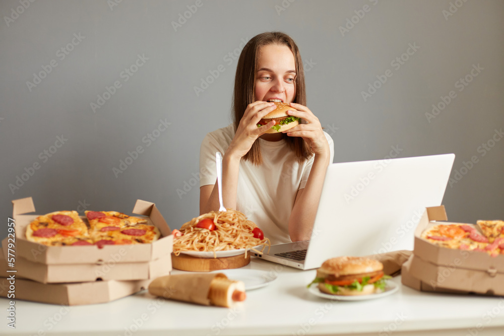 Portrait of dark haired attractive woman eating fast food, sitting at table against gray wall and watching film on notebook, biting big hamburger, having junk food snack. Portrait of dark haired attractive woman eating fast food, sitting at table against gray wall and watching film on notebook, biting big hamburger, having junk food snack.