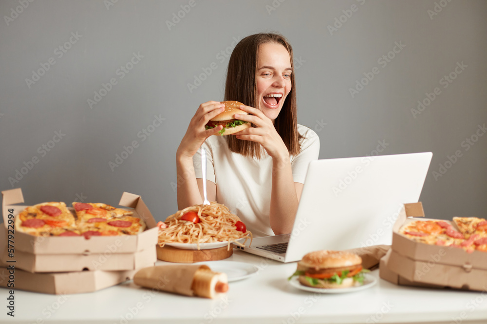 Image of satisfied delighted Caucasian woman eating fast food, sitting at table with lots junk food against gray wall, holding slice of pizza and hamburger in hands, keeps eyes closed. Image of satisfied delighted Caucasian woman eating fast food, sitting at table with lots junk food against gray wall, holding slice of pizza and hamburger in hands, keeps eyes closed.