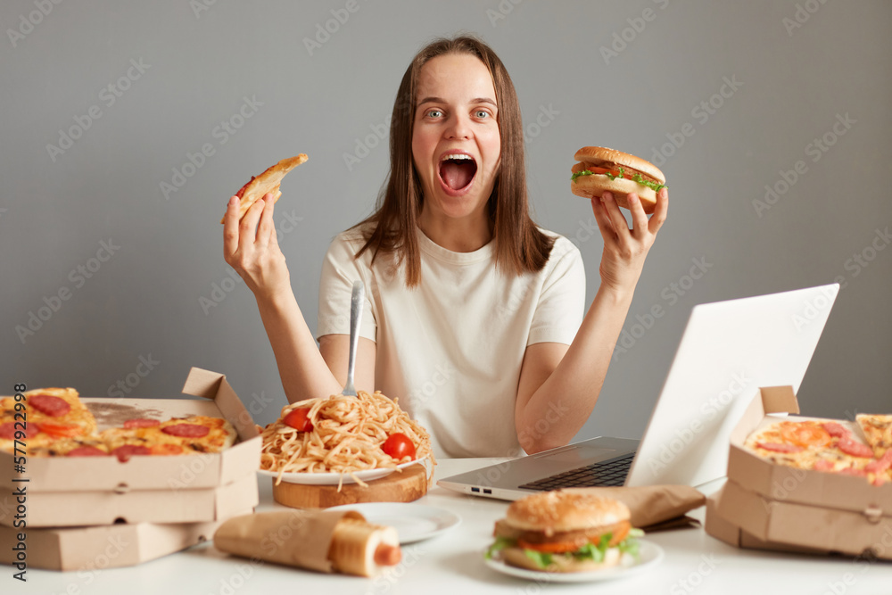 Portrait of excited young adult woman eating fast food while watching film on laptop, sitting at table against gray wall, holding sandwich and slice of pizza, screaming. Portrait of excited young adult woman eating fast food while watching film on laptop, sitting at table against gray wall, holding sandwich and slice of pizza, screaming.