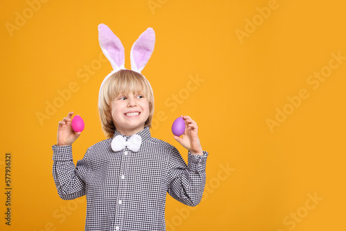 Happy boy in bunny ears headband holding painted Easter eggs on orange background. Space for text