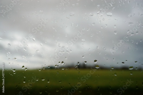Abstraktes Motiv mit Glasscheibe und Regentropfenmuster vor grünem Feld, Bäumen und grau-weißem Himmel bei Regen und Sturm am Morgen im Winter