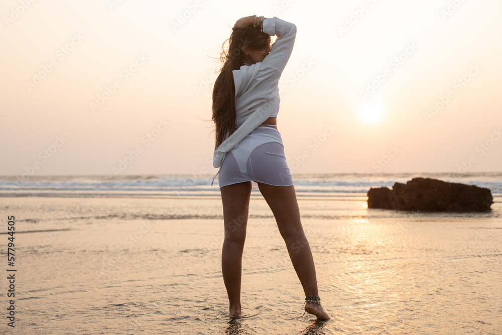 Tanned slender young woman with loose hair in white swimsuit stands with her back in sea water against sunset. 