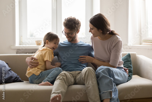 Happy young parents cuddling, tickling cheerful cute girl kid on home sofa, laughing, having fun, enjoying family leisure, parenthood. Positive couple playing with giggling little daughter on couch