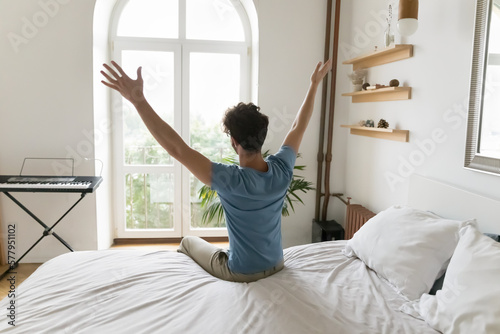 Positive excited young man enjoying good morning in cozy home bedroom, sitting on bed, feeling great after sleeping enough on comfortable mattress, rising hands, looking at window. Back view