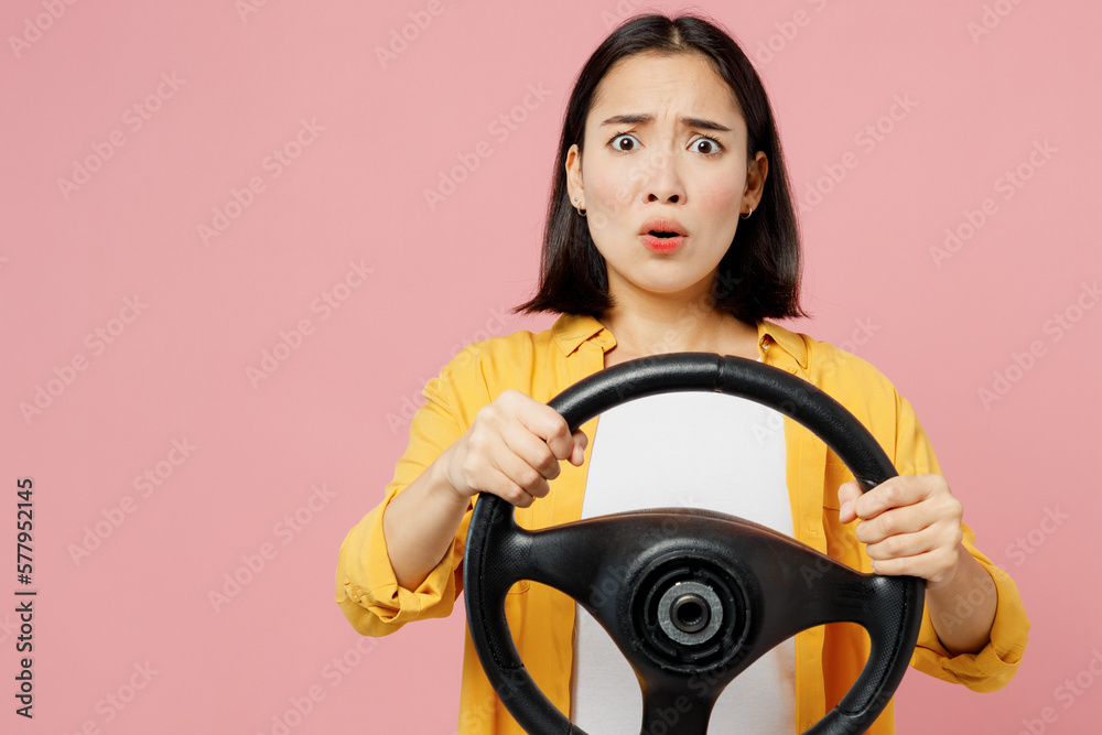 Young sad shocked woman of Asian ethnicity wear yellow shirt white t-shirt hold steering wheel driving car look aside on area isolated on plain pastel light pink background studio. Lifestyle concept. Young sad shocked woman of Asian ethnicity wear yellow shirt white t-shirt hold steering wheel driving car look aside on area isolated on plain pastel light pink background studio. Lifestyle concept.