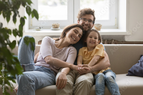 Cheerful attractive couple of young parents hugging cute girl on sofa, looking at camera, smiling, laughing. Family portrait of happy mom, dad and daughter kid enjoying closeness, leisure at home