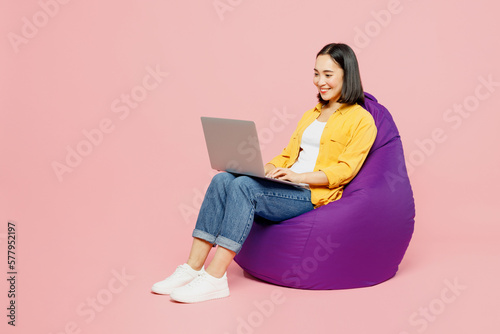 Full body young IT woman of Asian ethnicity wear yellow shirt white t-shirt sit in bag chair work hold use laptop pc computer isolated on plain pastel light pink background studio Lifestyle concept