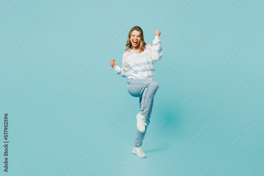 Full body overjoyed excited young woman wear striped hoody doing winner gesture celebrate clenching fists say yes isolated on plain pastel light blue cyan background studio portrait Lifestyle concept