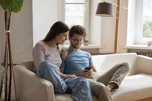 Happy young couple of husband and wife sitting on sofa together with gadget, using online app, media service on mobile phone, typing, looking at screen, laughing, smiling