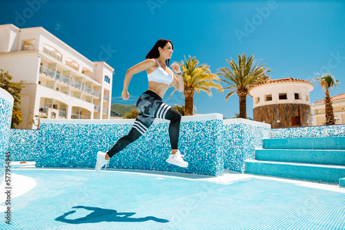 sporty woman running actively and jumping. Attractive fitness model jogging outdoors in sunny day. Swimming pool background. © shevtsovy