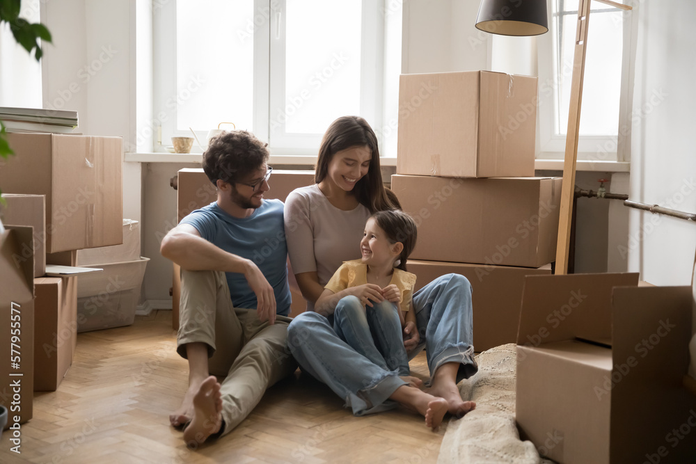Cheerful couple of parents and cute little daughter kid sitting on heating  floor of new home, apartment, resting among stacked cardboard boxes,  smiling, laughing, having fun, enjoying relocation Stock Photo