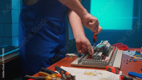Unrecognizable man in blue overalls with a screwdriver in his hands is screwing a red wire in an automatic electrical switch. Electrical workshop with tools on the table, blue light.