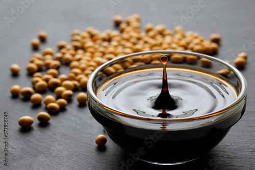 soy sauce with spalsh and drop in glass bowl and dry soybeans on black stone background