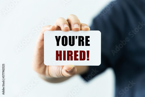 You're hired text on blank business card being held by a woman's hand with blurred background. Business concept to inform that you are hired.