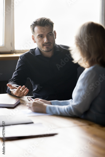 Millennial business professional man talking to female coworker at office table, speaking, explaining work project details, tasks, plan, strategy. Corporate mentor training intern