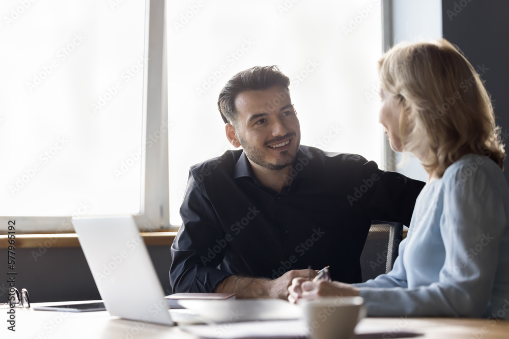 Couple of positive office coworkers chatting at workplace, talking at work desk with laptop, smiling, enjoying teamwork, cooperation. Business teacher mentoring intern, new employee