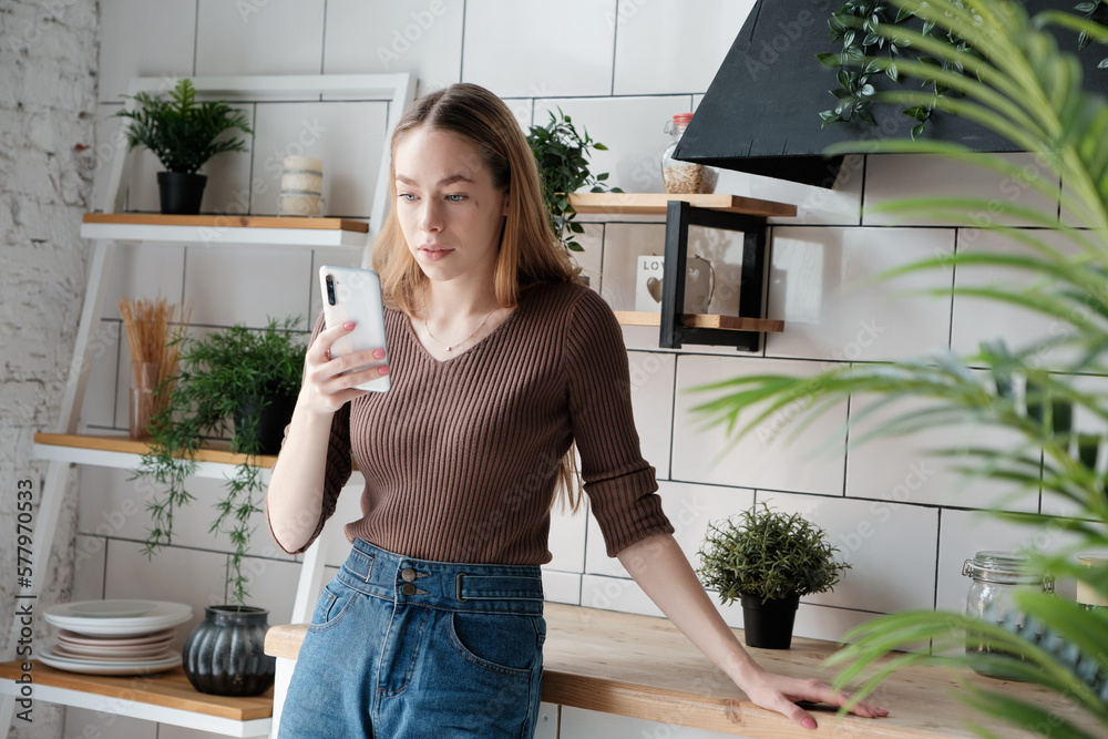 Young caucasian woman with long blonde hair using smartphone, typing writes a message, watches a video, reads a post in social networks at home in the kitchen with flowers in pots 