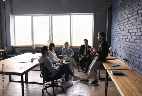 Focused millennial business group and team leader meeting in office loft interior, sitting in circle, listening to young male coach, mentor, trainer. Employees brainstorming on corporate briefing