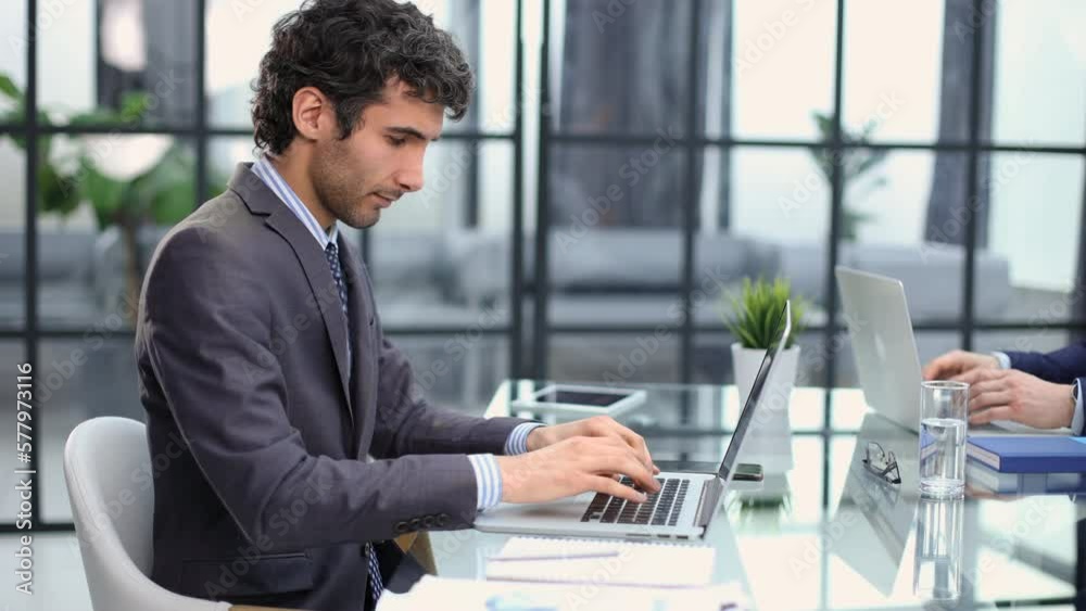 Portrait of a smiling young businessman working on laptop and smiling on camera at office with his colleague. Portrait of a smiling young businessman working on laptop and smiling on camera at office with his colleague.